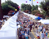 A view from 15 feet or so in the air of a large crowd of people meandering down a town street that is lined with balloons and white tents where artists are setup with booths and their work on display. A very festive feel with thousands of people enjoying sunny day.