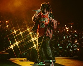 Rapper J. Cole performing on stage, wearing a Chicago Bulls jersey and large jacket, in front of a nighttime crowd with many holding phones and cameras in the background