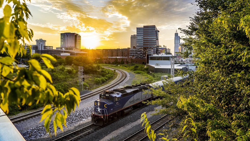 A colorful sunrise view of the Raleigh skyline from Boylan Bridge, with an Amtrak train in the foreground preparing to pass under the bridge 