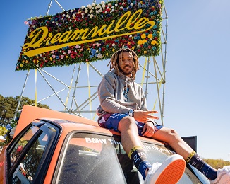 A man poses while sitting on top of an orange car in front of a large sign in the background that reads "Dreamville" at the Dreamville Festival at Dorothea Dix Park in Raleigh, N.C.