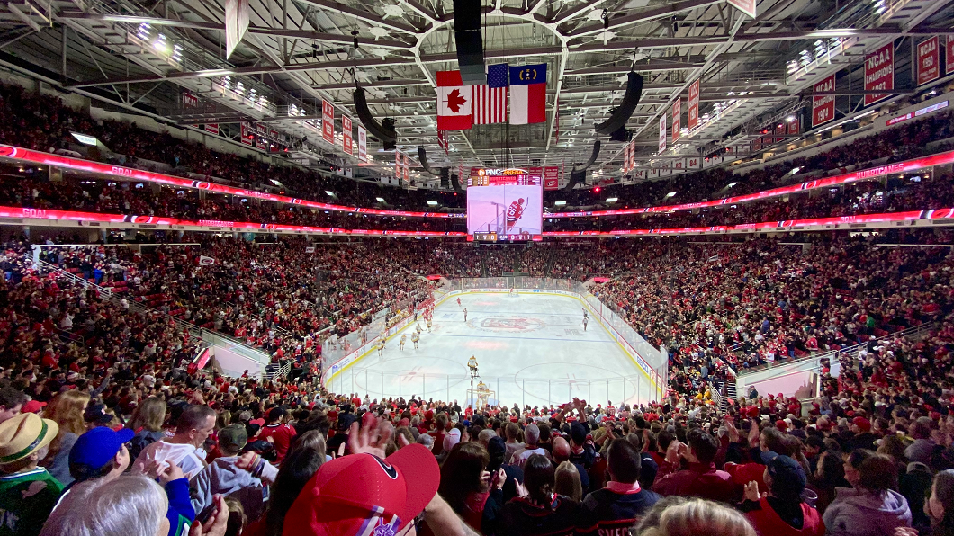 A packed crowd wearing mostly red colors fills PNC Arena for a Carolina Hurricanes hockey game; view is from behind one of the goals, about halfway up the arena