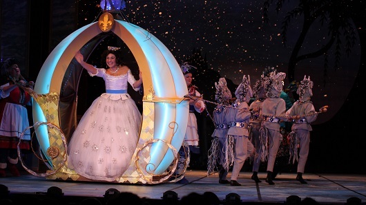 Photo of a theatre production of Cinderella | Children in horse costumes are pulling a pumpkin-shaped, round carriage as Cinderella waves to the audience