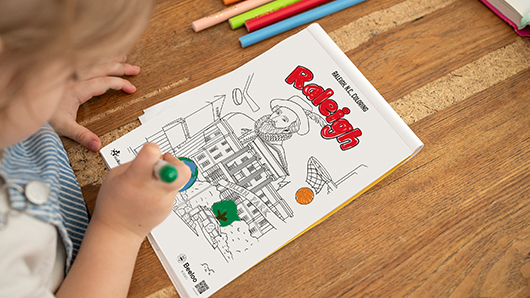 A view looking over a young girl's shoulder as she's coloring a Raleigh coloring sheet with red, green and orange markers and colored pencils; the coloring sheet is a collage of Raleigh-themed images, including Sir Walter Raleigh, the North Carolina State Capitol, a basketball hoop and ball and parks