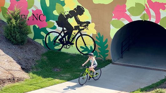 Woman riding a bike, coming out of a concrete tunnel with a giant bike mural painted on the exterior of the bridge