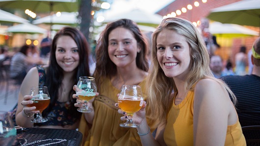 Three white woman in their 20s sit a table outdoors at a busy brewery, all holding half-empty tulip-style beer glasses and smiling at camera while leaning together