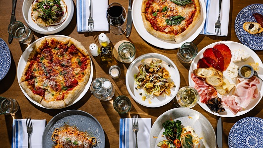 A top-down view of a full table of food at an upscale restaurant, with several plates of food that include pizza, pasta, charcuterie and more