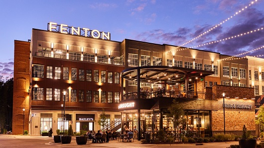 A dusk photo of the exterior of some buildings in a shopping center, with the word 'FENTON' displayed with lights on top of one of the buildings and a busy two-story bar in the foreground