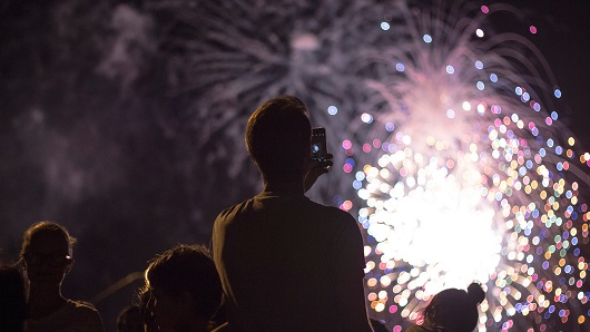 Crowd watches fireworks light up the night sky, man in foreground has back turned to camera while taking photo with his phone
