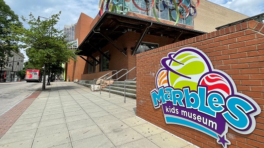 Exterior of Marbles Kids Museum, with a colorful 'Marbles' sign attached to a brick wall on the right, and the front entrance beyond that, with a large open sidewalk available