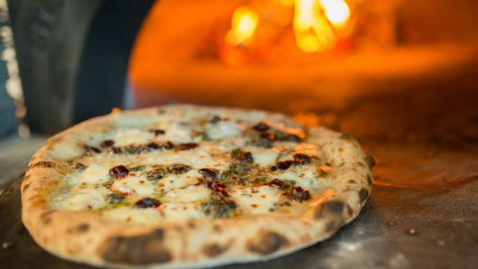 Photo of a cheesy pizza in front of a wood-fired grill