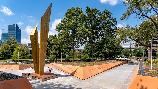 A wide, daytime view of the new North Carolina Freedom Park, with clay-colored walls lining concrete walkways that lead to the 45-foot-tall Beacon of Freedom in the center of the park