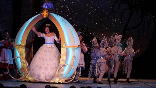 A view of the stage during a performance of "Cinderella," as Cinderella herself in a white ball gown waves out to the audience with a big smile as she's drawn by a carriage
