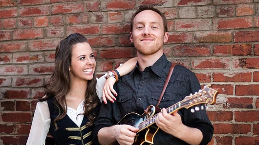 Artists Sierra Hull and Justin Moses smiling for posed photo in front of brick wall. Justin is holding a string instrument.