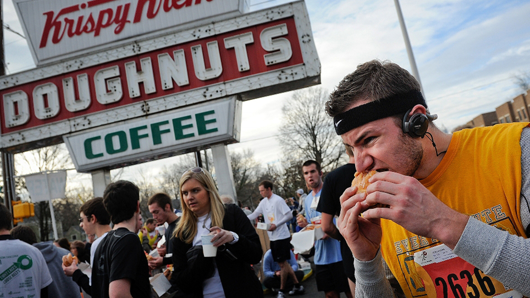 A group of runners scarfing down doughnuts in front of a Krispy Kreme Doughnuts and Coffee sign