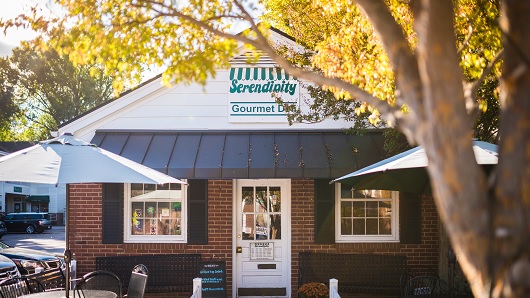 A view of the front door at Serendipity Gourmet Deli, a brick building with a black awning and white front door, plus a patio out front that has tables and a couple of umbrellas set up. The photo is taken during daytime with some yellow leaves on a tree out of focus near the camera.