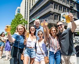 Group of six people holding beers hold them in the air while posing for group photo on a very sunny day in downtown Raleigh