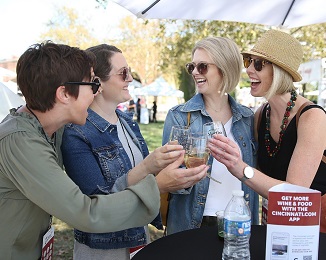 A group of four people at an outdoor event share a smile and laugh as they hold small wine glasses together 