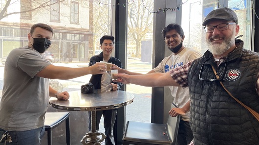 A group of four smile are smiling at the camera as they 'cheers' small coffee cups inside a coffee lounge that has large windows behind them. The group is on a coffee tasting tour of downtown Raleigh.