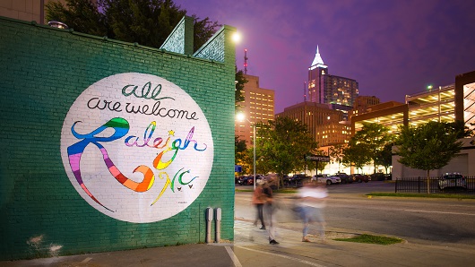 Slow-shutter photo of people walking past a mural that reads "Are All Welcome Raleigh, N.C." just a bit after sunset