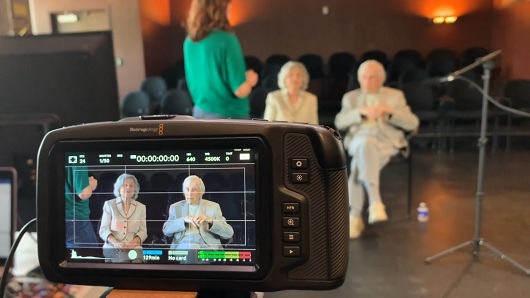 In the foreground is the back view of a camera that is recording video, within frame are two senior Jewish adults, a man and woman, who are preparing their microphones and getting ready to be interviewed; both people are wearing tan/beige outfits and have white hair; in the background a woman in a green top can be seen giving instructions