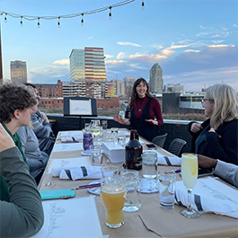 A woman instructing a watercolor art class; students are outside at a picnic table at a restaurant painting the downtown Raleigh skyline