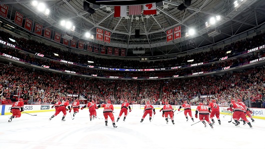 Group of a dozen hockey players wearing red jerseys skate together in a line away from the camera in front of packed crowd at indoor arena with bright lights overhead