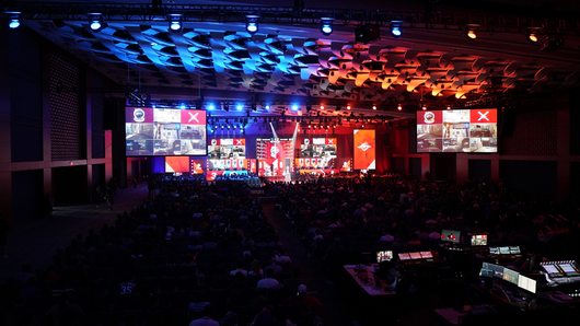 Interior view of a large esports event happening in an auditorium; a large crowd of spectators watch a stage where giant screens show player views in a multiplayer game