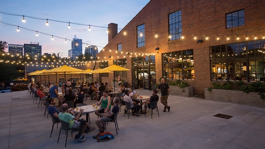 Dusk scene of the patio at Transfer Co. Food Hall, with string lights illuminating diners at tables with yellow umbrellas and the Raleigh Skyline shining in the background