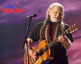 Willie Nelson on stage playing guitar in front of microphone, looking down at his left hand placement; background of the stage is a dark purple and pink color