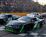 A black Chevrolet race car with green trim and the number seven on its side waits for a race at a checkered starting line, along with other cars in the background; packed spectator stands can be seen in the background under a clear evening sky