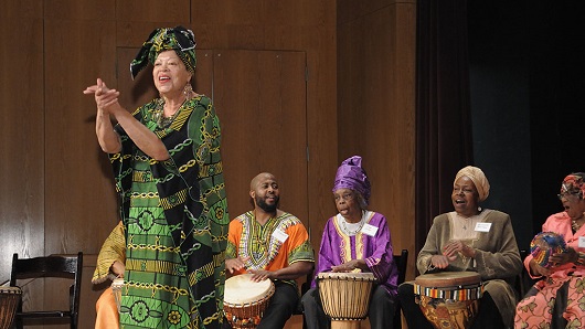 Black woman in traditional African American garment stands on stage clapping hands while several people sitting behind play drums with their hands 