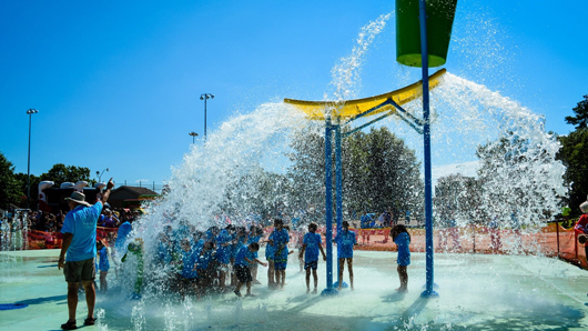 More than a dozen elementary aged children play in a splashpad with large buckets of water pouring down on the tightly bunched group