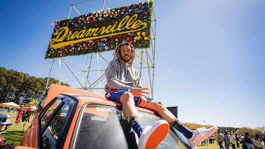 Festival attendee sits on hood or orange car on sunny day, looking at camera giving peace sign, with large sign in background that reads 'Dreamville'