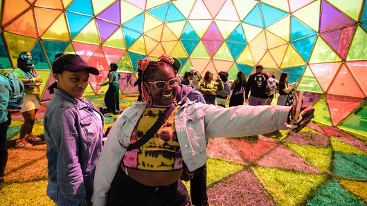 Two female Dreamville Festival attendees, both wearing denim jackets, pose for a selfie under a colorful globe at Dorothea Dix Park at a previous year's event