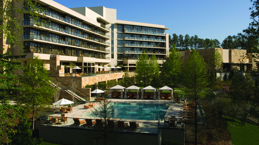 A view of The Umstead hotel's beautiful outdoor area, with cabanas setup pool-side, surrounded by tall trees and hotel room balconies looking over the area on a sunny day with blue skies