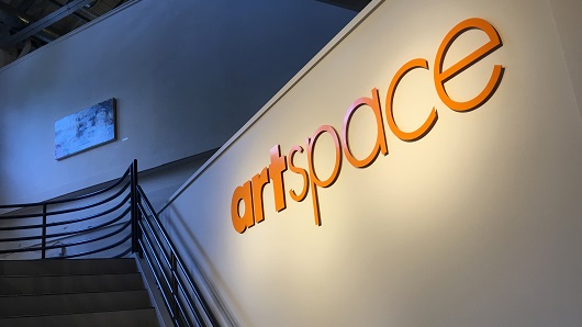 View of an interior wall at an art gallery and studio; word "artspace" in big, orange letters is on a white wall above a set of stairs