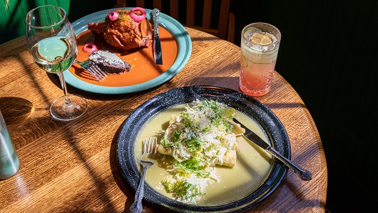 A 45-degree angle view of a restaurant table draped in sunlight with two dishes and two drinks; one plate has green chile chicken enchiladas and the other has Yucatan-style barbecued pork; drinks are a glass of white wine and a pinkish cocktail in a highball glass 