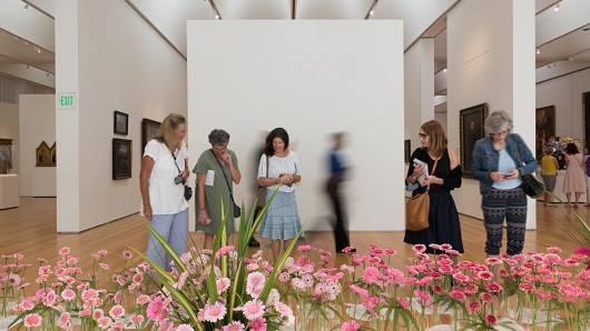 A group of women standing in an art gallery look down at a row of blooming pink flowers displayed in vases on the floor, filling the bottom half of the photo